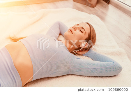 Top view portrait of relaxed woman listening to music with headphones lying on carpet at home. She is dressed in a blue tracksuit. 109860000