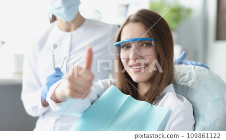 Young woman in safety glasses sitting in dental chair and showing thumbs up Young woman in safety glasses sitting in dental chair and showing thumbs up 109861122
