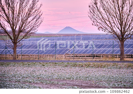 Solar power plant park with panels beyond chain link fence. Subsidized renewable energy source on arable land. Europe. Czech Republic. Freezing winter morning or evening in agricultural field. 109862462