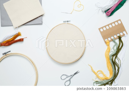 Embroidery set fot stitching. Beige cotton cloth in embroidery hoop on white background with fabric, colorful threads, scissors and needls. Indoor hobby concept with copy space, top view Embroidery set fot stitching. Beige cotton cloth in embroidery hoop on white background with fabric, colorful threads, scissors and needls. Indoor hobby concept with copy space, top view 109863863