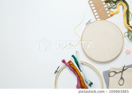 Embroidery set fot stitching. Beige cotton cloth in embroidery hoop on white background with fabric, colorful threads, scissors and needls. Indoor hobby concept with copy space, top view 109863865
