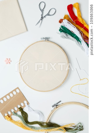 Embroidery set fot stitching. Beige cotton cloth in embroidery hoop on white background with fabric, colorful threads, scissors and needls. Indoor hobby concept with copy space, top view Embroidery set fot stitching. Beige cotton cloth in embroidery hoop on white background with fabric, colorful threads, scissors and needls. Indoor hobby concept with copy space, top view 109863866