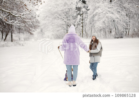 Mother, daughter and their dog french bulldog walking in winter on a snow. Woman and girl wearing warm jackets and knitted hat. Dog wearing warm overalls. 109865128