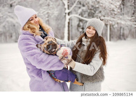 Mother, daughter and their dog french bulldog playing in winter on a snow. Woman and girl wearing warm jackets and knitted hat. Dog wearing warm overalls. 109865134