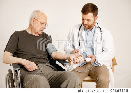 Senior gentleman on a wheelchair isolated on a white background. Man wearing khaki t-shirt and doctor wearing white medical gown. Doctor talking with man and measured his arterial pressure. 109865166