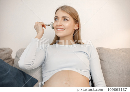 Portrait of pregnant woman holding a face massager. Pregnant woman sitting on a sofa in living room. Blonde woman wearing beige jacket. Portrait of pregnant woman holding a face massager. Pregnant woman sitting on a sofa in living room. Blonde woman wearing beige jacket. 109865187