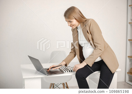 Portrait of pregnant woman typing on a laptop while sitting on desk in modern office. Pregnant businesswoman using a laptop while sitting in her office. Blonde woman wearing beige jacket. 109865198