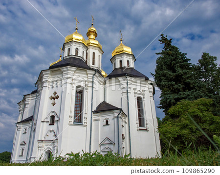 The Orthodox Church of St. Catherine in Chernihiv, with its white facade, iconic golden domes, and crosses, harmonizing beautifully with the vibrant greenery and the serene blue sky. 109865290