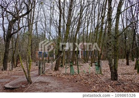 A structure with a blue blue windows surrounded by trees. The facade of an abandoned kindergarten in the Chernobyl radioactive zone. Autumn foliage on the ground. Trees in front of an old house. A structure with a blue blue windows surrounded by trees. The facade of an abandoned kindergarten in the Chernobyl radioactive zone. Autumn foliage on the ground. Trees in front of an old house. 109865326