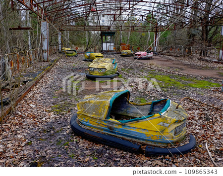 A rusted, abandoned yellow bumper car with empty seats surrounded by overgrown vegetation. 109865343