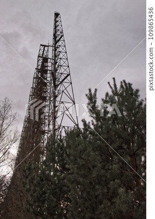 The image shows a tall, rusted metal tower standing amidst green trees under a cloudy sky. Duga is a Soviet over-the-horizon radar station for an early detection system for ICBM launches. The image shows a tall, rusted metal tower standing amidst green trees under a cloudy sky. Duga is a Soviet over-the-horizon radar station for an early detection system for ICBM launches. 109865436