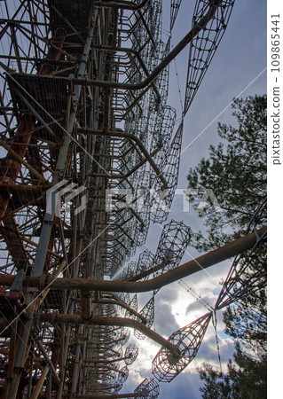 A complex structure of rusted metal beams and wire grids against a cloudy sky, surrounded by trees. Duga is a Soviet over-the-horizon radar station for an early detection system for ICBM launches. A complex structure of rusted metal beams and wire grids against a cloudy sky, surrounded by trees. Duga is a Soviet over-the-horizon radar station for an early detection system for ICBM launches. 109865441