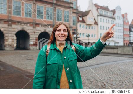 Happy cheerful young woman on city street. Redhead girl with backpack pointing finger on urban background. Traveling Europe in autumn. Urban lifestyle concept. Happy cheerful young woman on city street. Redhead girl with backpack pointing finger on urban background. Traveling Europe in autumn. Urban lifestyle concept. 109866328