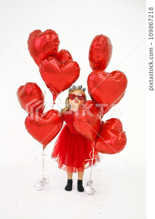 A funny girl in a red dress and heart-shaped sunglasses hides between balloons and blows a kiss A funny girl in a red dress and heart-shaped sunglasses hides between balloons and blows a kiss 109867218