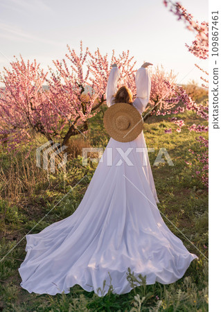 Woman blooming peach orchard. Against the backdrop of a picturesque peach orchard, a woman in a long white dress and hat enjoys a peaceful walk in the park, surrounded by the beauty of nature. Woman blooming peach orchard. Against the backdrop of a picturesque peach orchard, a woman in a long white dress and hat enjoys a peaceful walk in the park, surrounded by the beauty of nature. 109867461