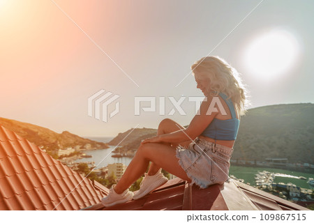 Woman sits on rooftop, enjoys town view and sea mountains. Peaceful rooftop relaxation. Below her, there is a town with several boats visible in the water. Rooftop vantage point. Woman sits on rooftop, enjoys town view and sea mountains. Peaceful rooftop relaxation. Below her, there is a town with several boats visible in the water. Rooftop vantage point. 109867515