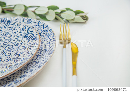 On the side, two serving plates with blue patterns on a white background stand one on one, part of the plates can be seen, gold-white appliances are located on the side, a green branch of plants lies 109868471