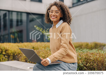 Cheerful woman manager in eyeglasses is working on laptop sitting on background of office building Cheerful woman manager in eyeglasses is working on laptop sitting on background of office building 109870075