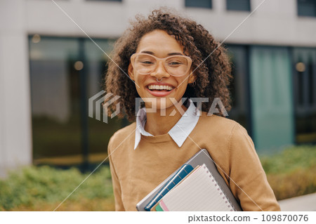 Young smiling female student standing with laptop and note pads on modern building backgrounds Young smiling female student standing with laptop and note pads on modern building backgrounds 109870076