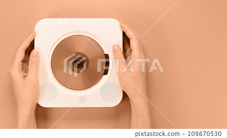 Hands holding stylish white compact CD player. Image toned in Peach Fuzz color of the year 2024. Retro vintage trending disc player. Top view, space for text. 109870530