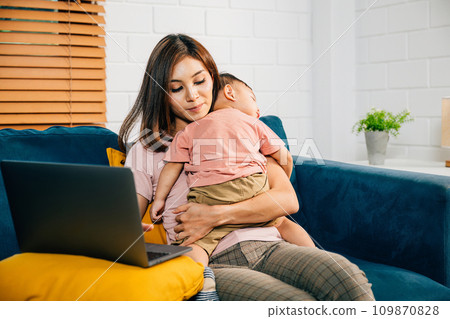 Balancing motherhood and work a businesswoman types on her laptop as her baby daughter peacefully naps on the sofa. This portrait captures their loving bond. 109870828