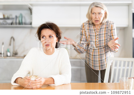 Upset middle-aged woman sitting at the kitchen table with her back to old woman quarreling to her 109872951