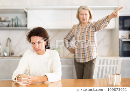 Upset middle-aged woman sitting at the kitchen table with her back to old woman quarreling to her Upset middle-aged woman sitting at the kitchen table with her back to old woman quarreling to her 109873031