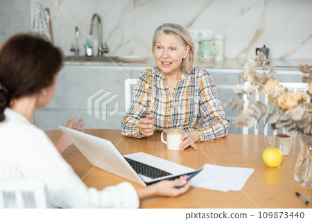 Positive old woman sitting at the kitchen-table face to face with middle-aged female agent Positive old woman sitting at the kitchen-table face to face with middle-aged female agent 109873440