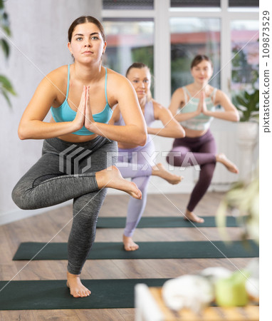 Diligent women practicing tree pose of yoga in light fitness room Diligent women practicing tree pose of yoga in light fitness room 109873529