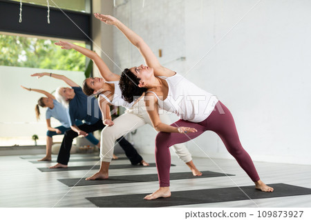 Concentrated young woman performing Revolved Side Angle Pose Parivrtta Parsvakonasana during yoga class with female group in fitness studio 109873927