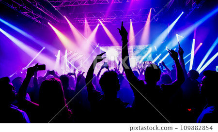 People dancing at party with hands up. Colorful concert crowd in front of lit stage inside concert venue, during music festival. Colorful laser show in club 109882854