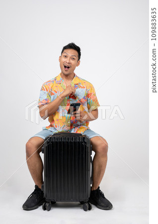 A fun, excited Asian man in summer clothes is sitting on his luggage on an isolated white background 109883635