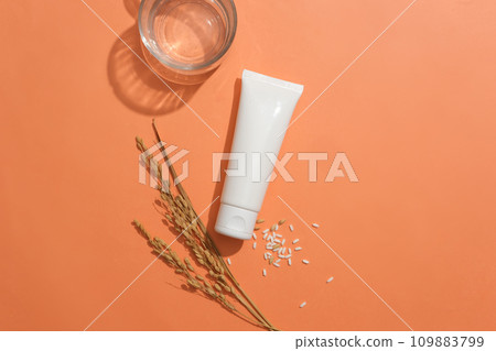 Centered unlabeled white tube with white rice and a glass on an orange background. Embracing a vegan cosmetics concept featuring rice extract. Ample copy space. Centered unlabeled white tube with white rice and a glass on an orange background. Embracing a vegan cosmetics concept featuring rice extract. Ample copy space. 109883799