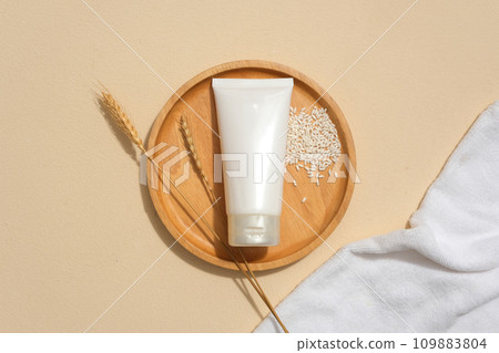 Flat lay of a tube of facial cleanser displayed on a round wooden plate with whole grain rice. Blank labels for branding and advertising. Minimalist background. 109883804