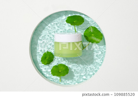 A jar of green lotion and pennywort leaves are placed on a glass platform with a ripple effect. Blank label for lettering design. Space for advertising. 109885520