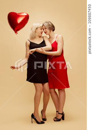 Happy young women, lesbian couple in elegant dresses celebrating romantic holiday over beige background. Valentine's Day, holiday, lgbt Happy young women, lesbian couple in elegant dresses celebrating romantic holiday over beige background. Valentine's Day, holiday, lgbt 109887450
