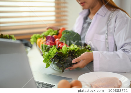 Nutritionist holding glass tray of fresh vegetables promoting a healthy diet during online consultation. 109888751