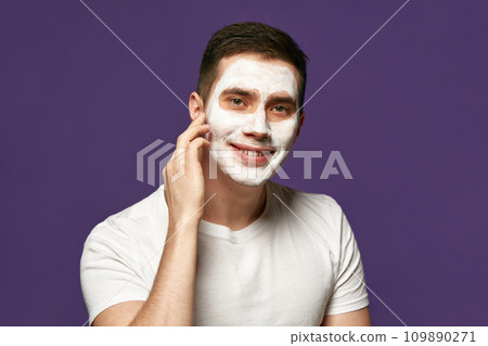 Portrait of calm brunette man, attractive guy with facial care mask, cosmetic product against purple background. 109890271