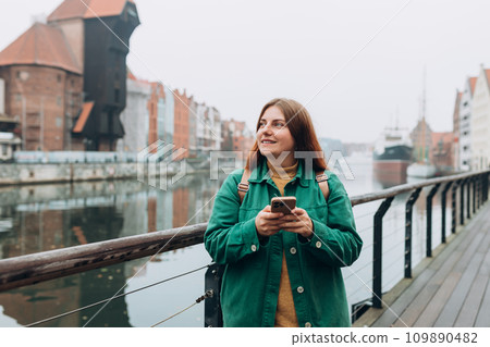 Attractive young female tourist is exploring new city. Gdansk old town and famous Zuraw crane, A happy beautiful 30s woman with smart phone is standing near Motlawa river Attractive young female tourist is exploring new city. Gdansk old town and famous Zuraw crane, A happy beautiful 30s woman with smart phone is standing near Motlawa river 109890482