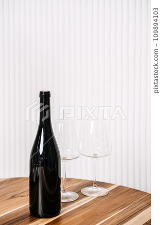 Minimalist Wine Bottle and Glasses on Wooden Table. A simple yet elegant setup of a wine bottle and two glasses on a polished wooden table against a striped background. 109894103