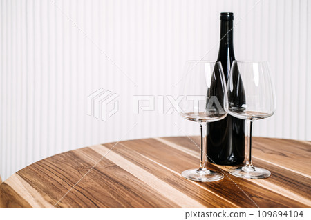 Minimalist Wine Bottle and Glasses on Wooden Table. A simple yet elegant setup of a wine bottle and two glasses on a polished wooden table against a striped background. Minimalist Wine Bottle and Glasses on Wooden Table. A simple yet elegant setup of a wine bottle and two glasses on a polished wooden table against a striped background. 109894104