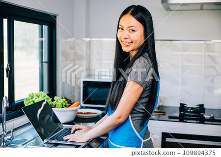 In the kitchen a smiling woman stands engrossed in cooking and using her laptop. Emphasizing the joy of technology in aiding culinary experiments and learning new recipes. 109894375