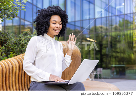 Young African American businesswoman working on laptop, talking on video call online, greeting and waving at camera. Sitting on a bench outside an office building. Young African American businesswoman working on laptop, talking on video call online, greeting and waving at camera. Sitting on a bench outside an office building. 109895377