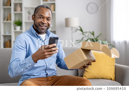 Portrait of an African-American man sitting on the sofa at home, holding an open parcel box in his hand, using a mobile phone, looking and smiling at the camera. 109895410