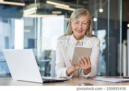 Woman boss at workplace inside office, senior experienced gray-haired business woman uses tablet computer, happy checking financial data, banker finance woman with laptop. Woman boss at workplace inside office, senior experienced gray-haired business woman uses tablet computer, happy checking financial data, banker finance woman with laptop. 109895436