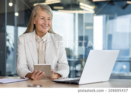 Woman boss at workplace inside office, senior experienced gray-haired business woman uses tablet computer, happy checking financial data, banker finance woman with laptop. Woman boss at workplace inside office, senior experienced gray-haired business woman uses tablet computer, happy checking financial data, banker finance woman with laptop. 109895437