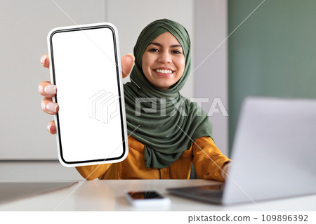 Smiling arab woman sitting at workplace and demonstrating blank smartphone at camera Smiling arab woman sitting at workplace and demonstrating blank smartphone at camera 109896392