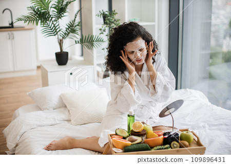 Lady fixing facial mask using cosmetic mirror on bed indoors Lady fixing facial mask using cosmetic mirror on bed indoors 109896431