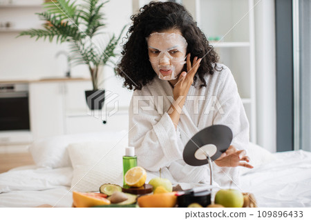 Female in bathrobe putting tissue mask on face in bedroom Female in bathrobe putting tissue mask on face in bedroom 109896433