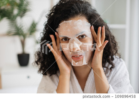 Woman in after-shower outfit touching cosmetic mask fixed on face Woman in after-shower outfit touching cosmetic mask fixed on face 109896458